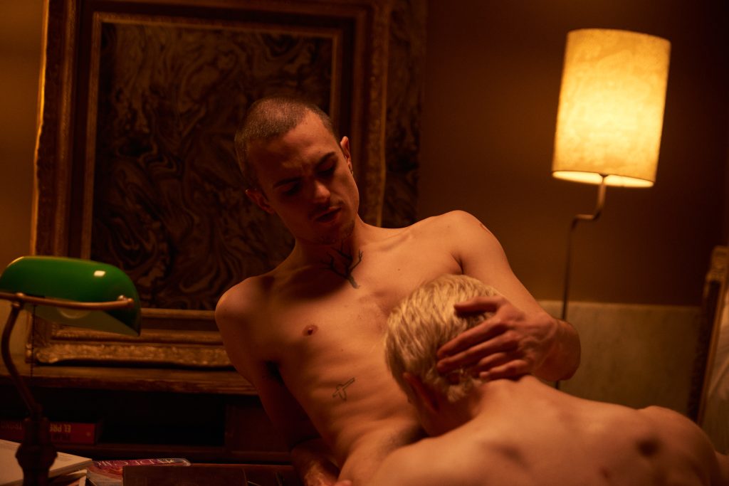 Two men engaged in a sensual and explicit encounter during a ritual gathering.